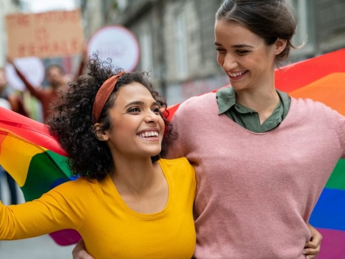 ragazze che si abbracciano sotto la bandiera del gay pride, uso della schwa